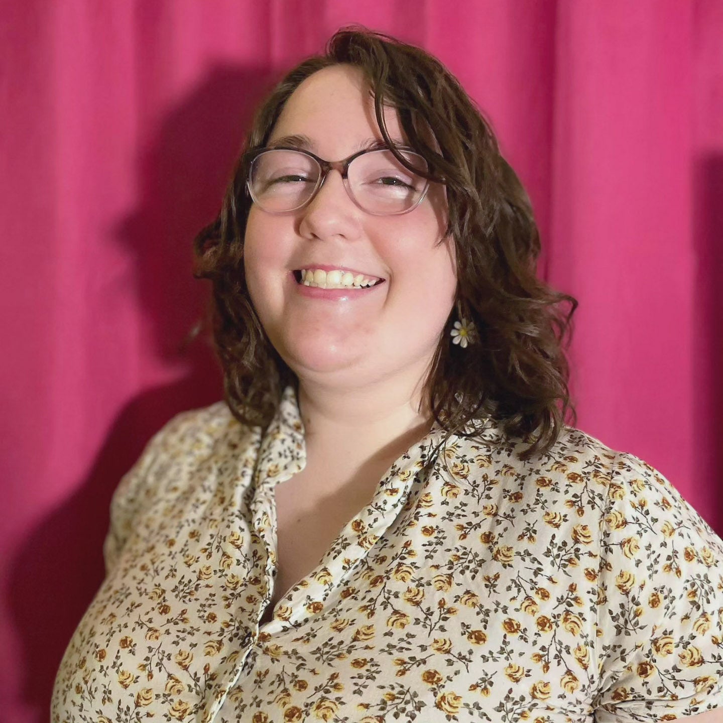 A young woman with brown curly hair wearing a floral shirt smiles brightly.