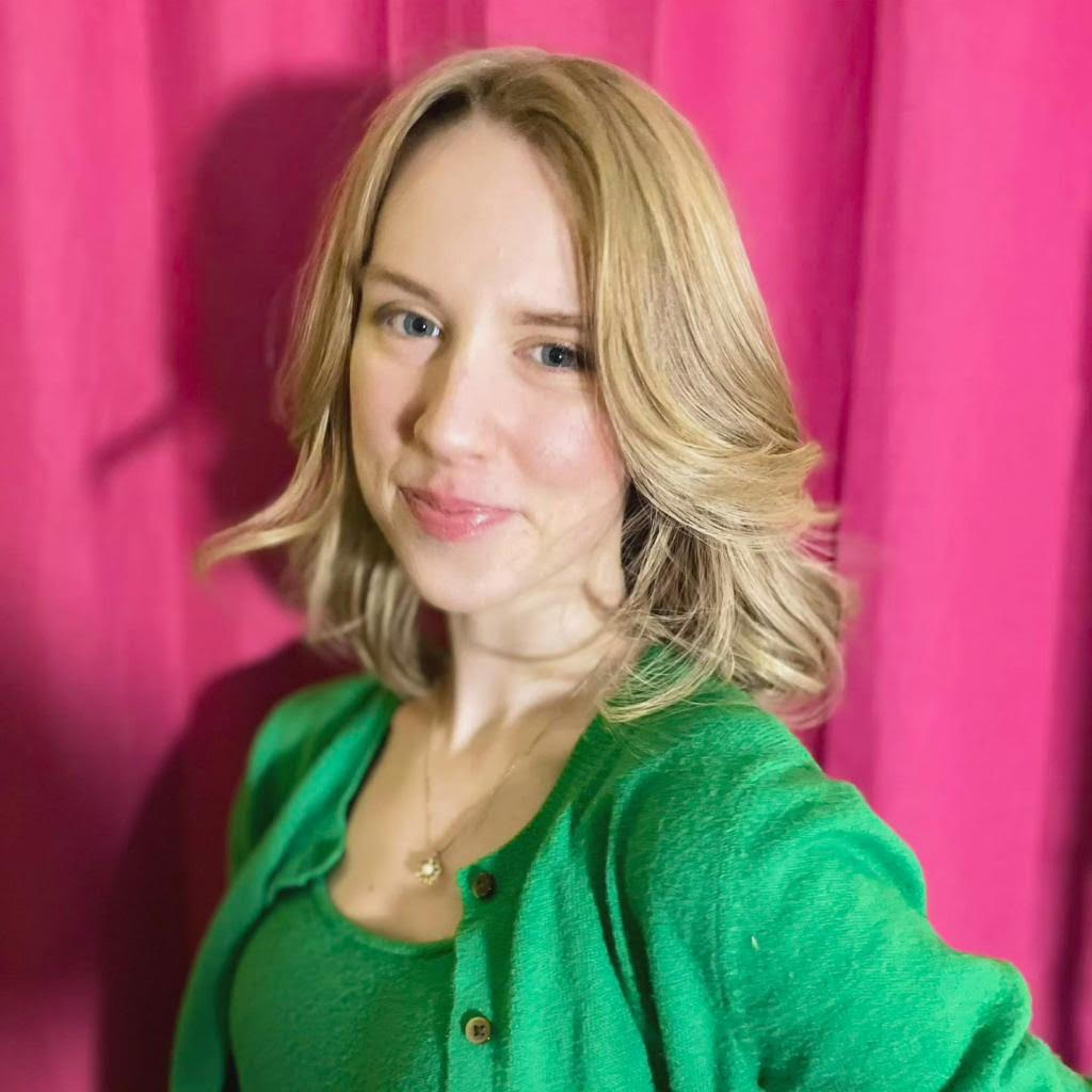 A young white woman with shoulder length blonde hair in a green outfit smiles sweetly. 