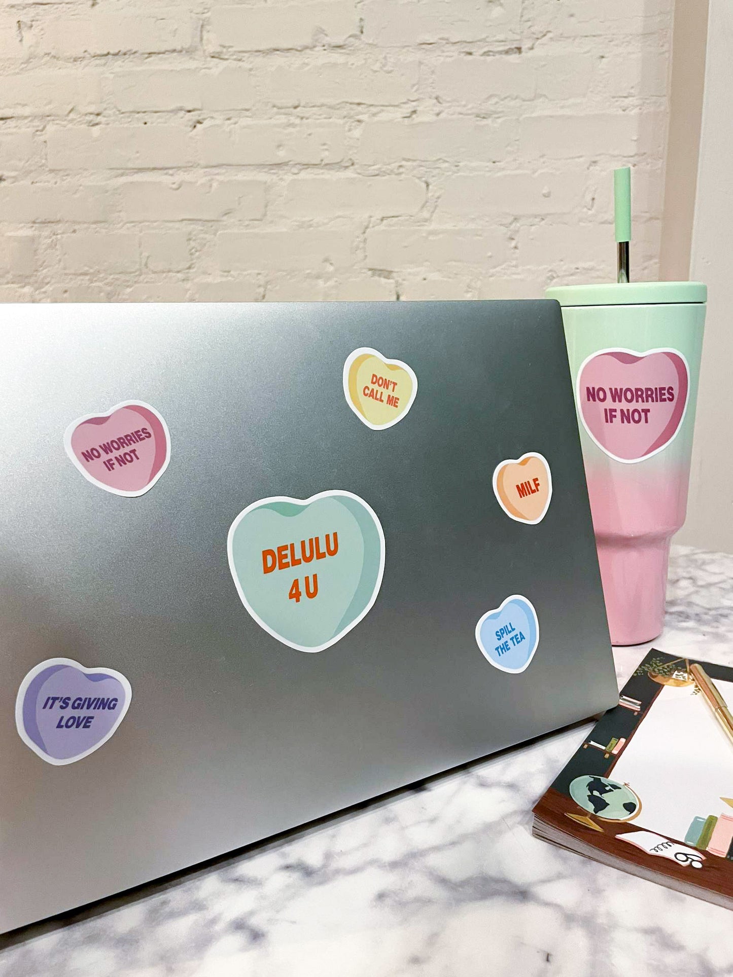 Laptop with five small candy heart stickers in different colors, plus a large green one that reads "delulu for you." A water bottle next to the laptop has a pink conversation heart sticker that reads "no worries if not."